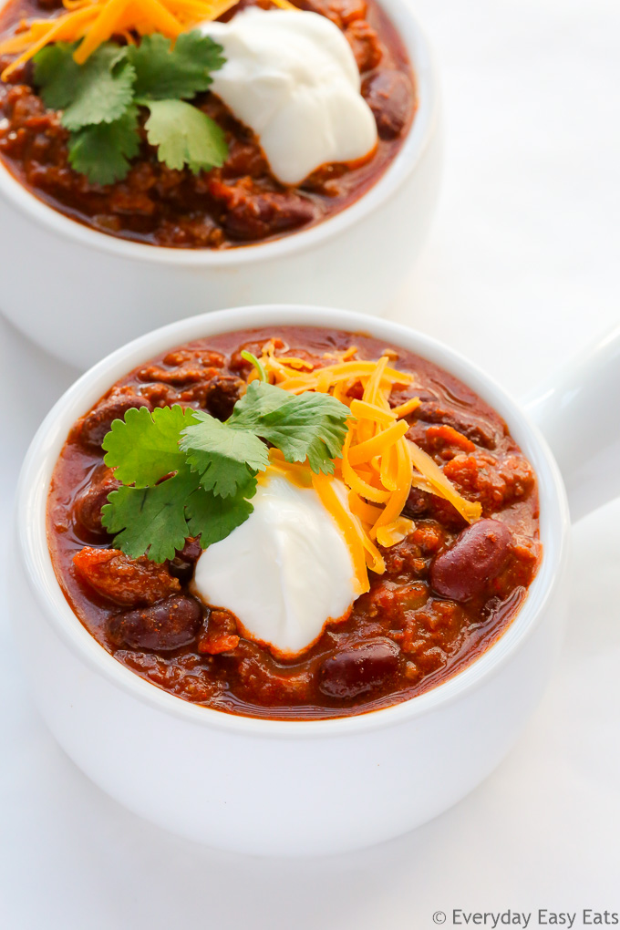 Healthy Meat Recipe: Overhead view of two bowls of Ground Beef Chili, garnished with shredded cheese, cilantro and sour cream, on a white surface.